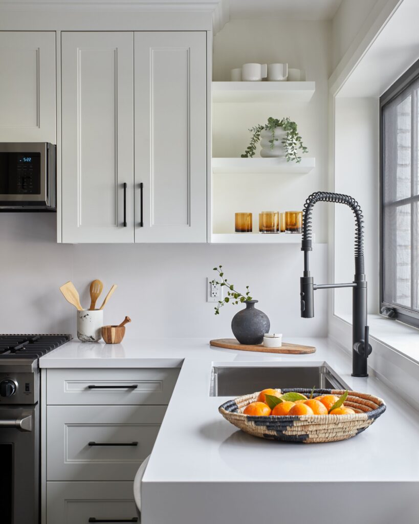 Modern white kitchen with black fixtures, open shelves with glassware and plants, a bowl of fresh citrus fruit on the counter, and sunlight streaming through a large window by the sink.
