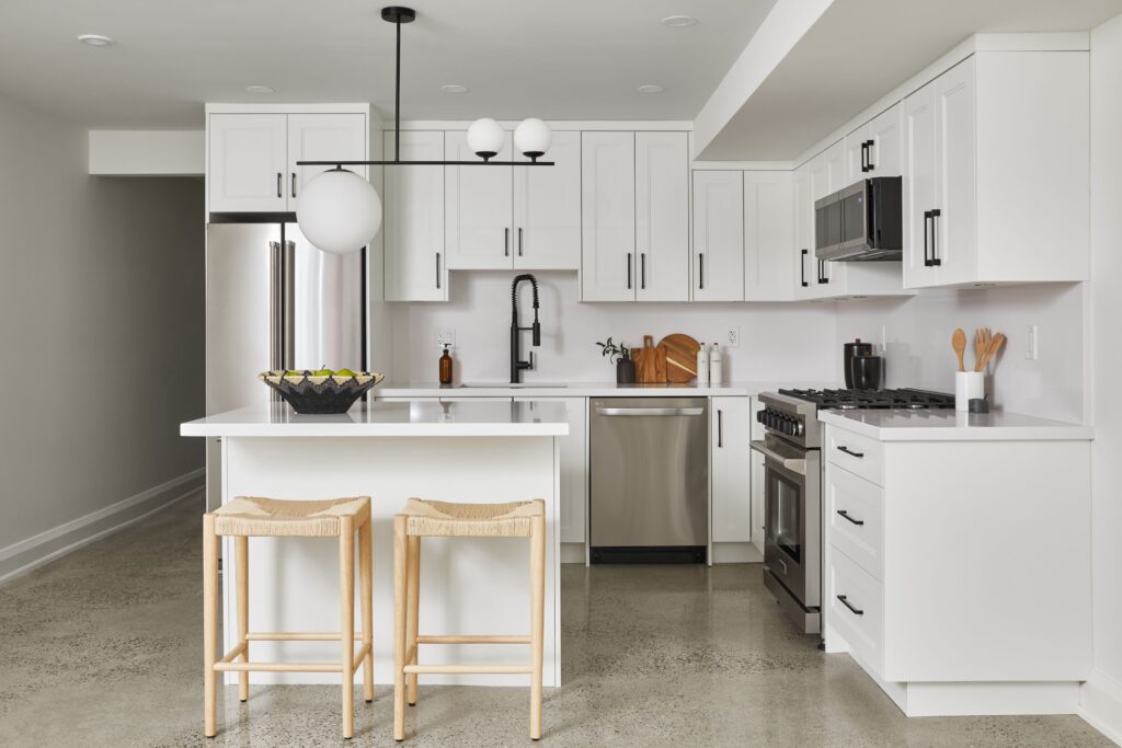 Modern kitchen with white cabinets, stainless steel appliances, a central island with two wooden stools, black faucet, fruit bowl, and minimal décor. Pendant light fixture hangs above the island.
