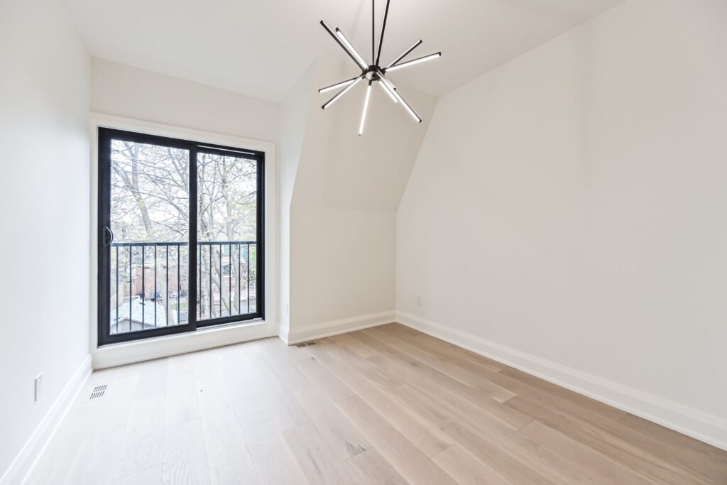 A bright, empty room with light wood floors, white walls, a modern black chandelier, and large glass doors leading to a balcony with a view of trees outside.