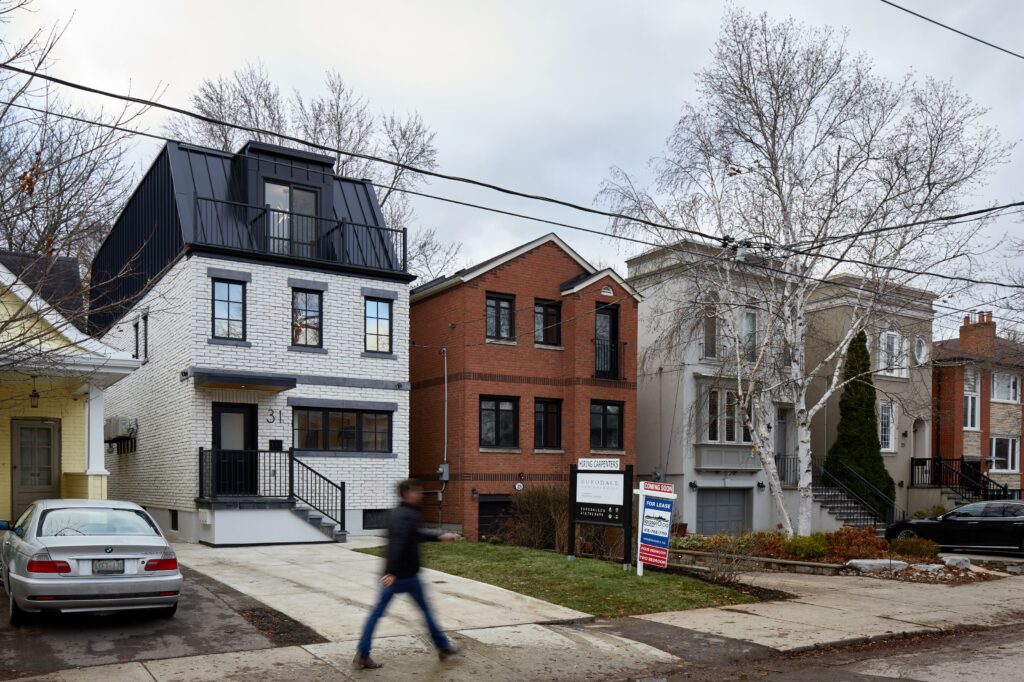 A person walks past modern two- and three-story houses on a residential street, with a for sale sign posted in front of one home. It is overcast, and trees are leafless, suggesting fall or winter.