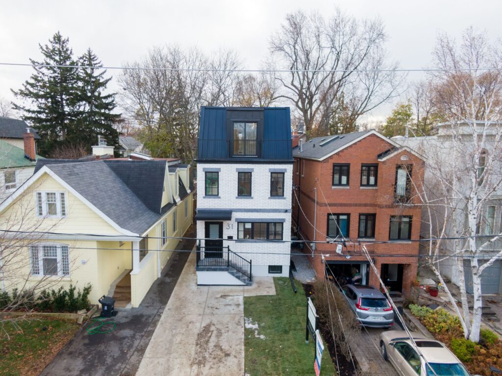 A modern three-story house with a dark upper floor and white brick lower floors stands between a yellow house and a red brick house. Cars are parked in the driveways and trees without leaves line the street.