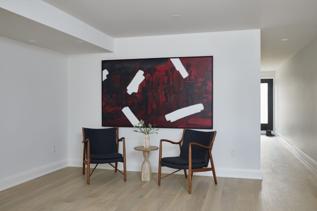 A modern, minimal room with two dark chairs and a small round side table holding a vase of flowers, set against a white wall featuring a large red abstract artwork. Light wood flooring and an open hallway are also visible.