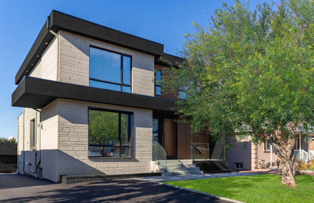 Modern two-story house with large windows, light-colored brick exterior, black trim, flat roof, and a front yard featuring a green lawn and a leafy tree under a clear blue sky.
