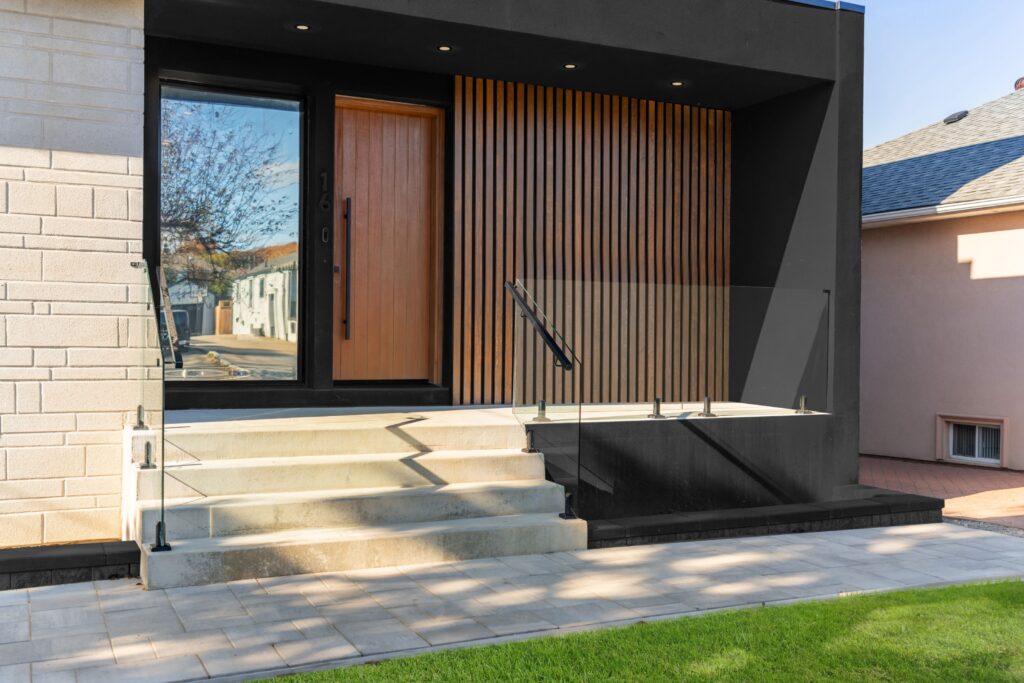 A modern house entrance with a wooden door, vertical wooden slats on the wall, glass railings, and concrete steps leading up from a stone pathway, with green grass in the foreground.