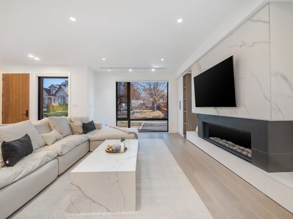 Modern living room with a large white sectional sofa, marble coffee table, wall-mounted TV above a sleek fireplace, and floor-to-ceiling windows showing a suburban view. Room features neutral colors and wood accents.