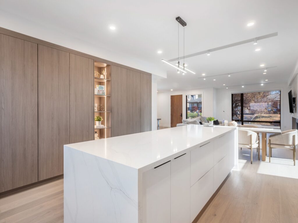 Modern kitchen with a large white marble island, minimalist white cabinets, light wood floors, built-in shelving with decor, recessed lighting, and large windows letting in natural light.