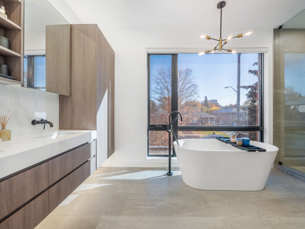 Modern bathroom with a freestanding white tub near large windows, wood cabinets, a long counter with sink, and a contemporary light fixture. Sunlight fills the space, highlighting neutral tones and clean lines.