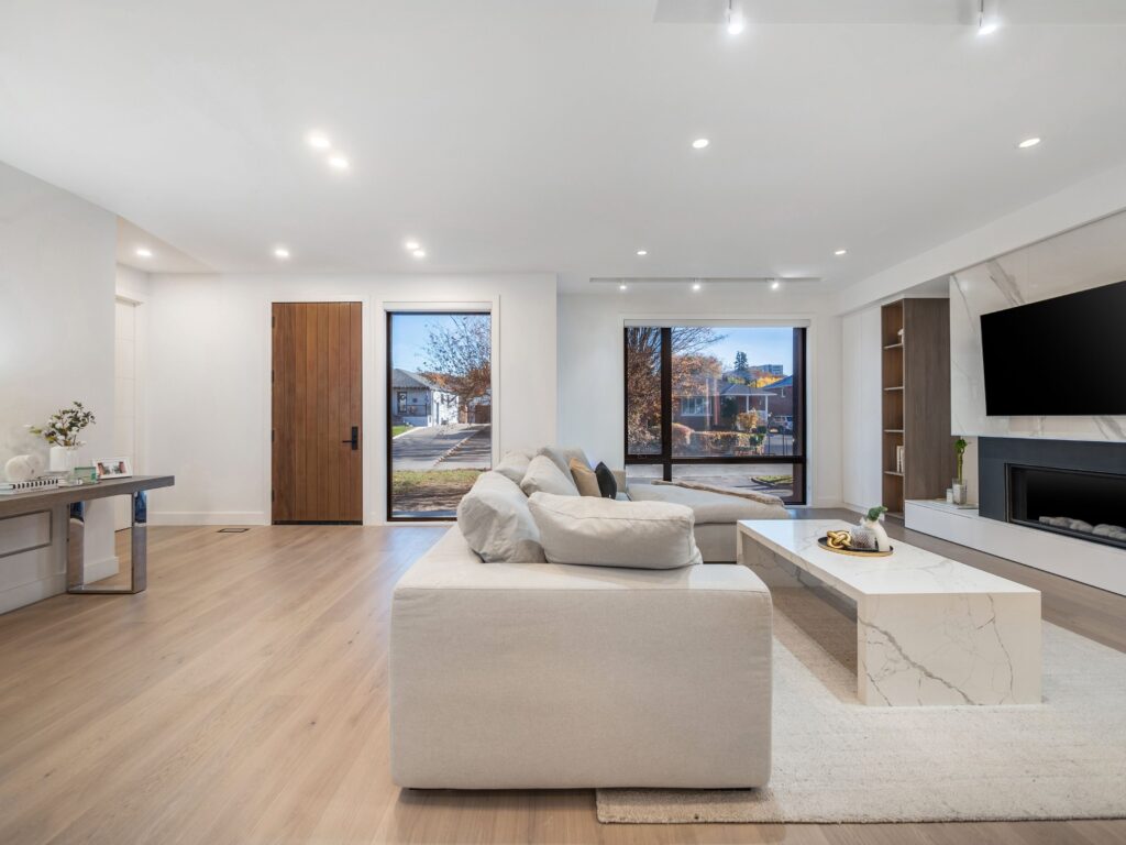 Modern living room with light wood floors, a white sectional sofa, marble coffee table, wall-mounted TV above a fireplace, large windows, and a wooden front door letting in natural light.