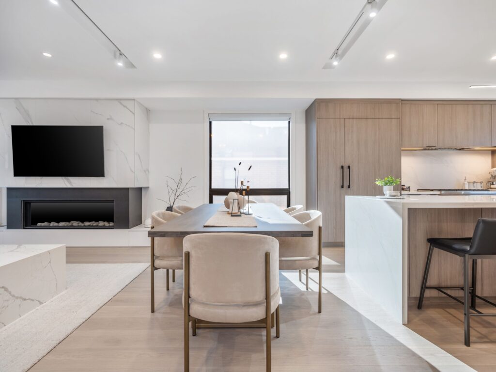 Modern open-concept kitchen and dining area with light wood flooring, beige chairs around a wooden table, marble countertops, built-in fireplace, wall-mounted TV, and minimalistic decor.