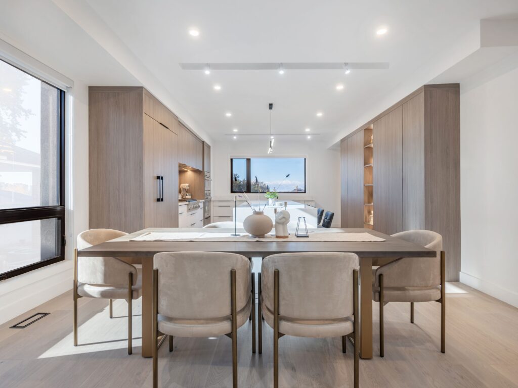 A modern dining room with a wooden table, six upholstered chairs, minimalist decor, and built-in wood cabinetry. Large windows and recessed lighting brighten the neutral-toned space. A kitchen is visible in the background.