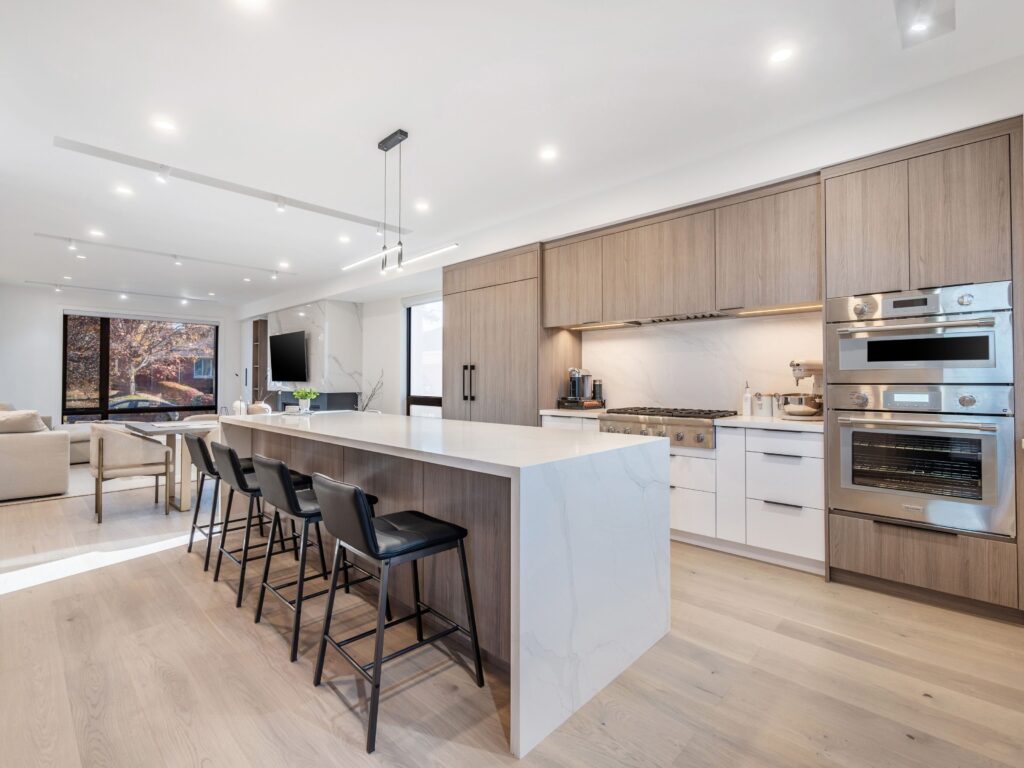 Modern kitchen with light wood floors, large marble island with four black chairs, built-in stainless steel appliances, light wood cabinets, and open living area with dining table and couch near large windows.