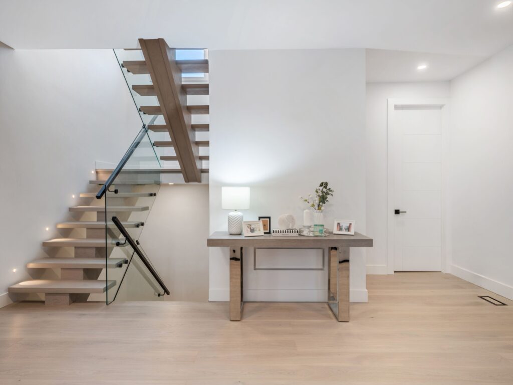 A modern, minimalist entryway with light wood floors, a glass-railed floating staircase, and a wooden console table displaying decor, photos, and a lamp, all against white walls.