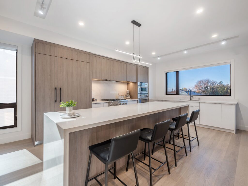 Modern kitchen with light wood cabinets, white countertops, a large island with four black chairs, built-in appliances, under-cabinet lighting, and a large window with a view of trees. A small plant sits on the island.