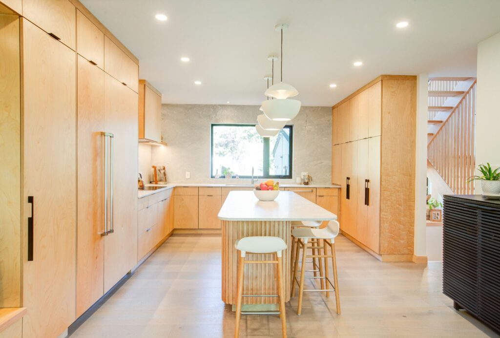 Modern kitchen with light wood cabinets, a large central island with stools, pendant lights above, and a window over the sink. The space is bright, open, and minimalist with neutral tones and natural lighting.