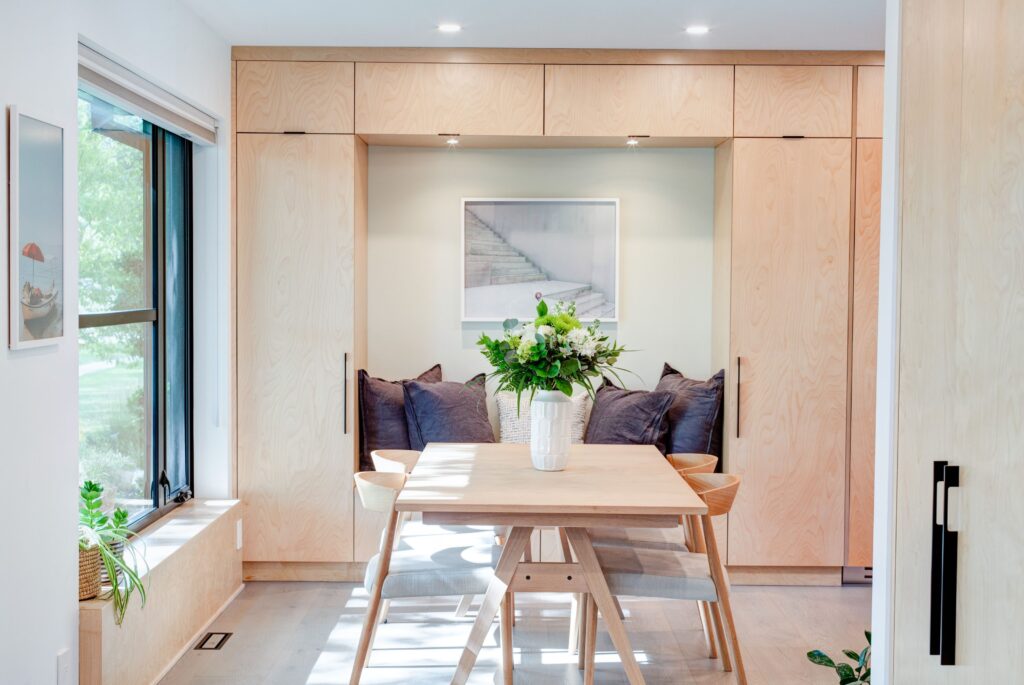 A modern dining nook with light wood cabinetry, a small wooden table, two chairs, gray cushions, and a large vase of flowers. A framed photo and a window with greenery are visible in the bright, airy room.