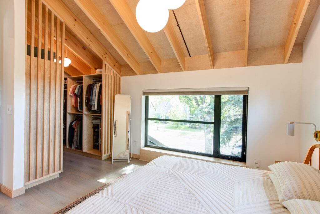 A modern bedroom with a wood-beamed ceiling, a large window letting in natural light, a bed with a white quilt, a wall-mounted lamp, an open closet with hanging clothes, and a tall mirror.