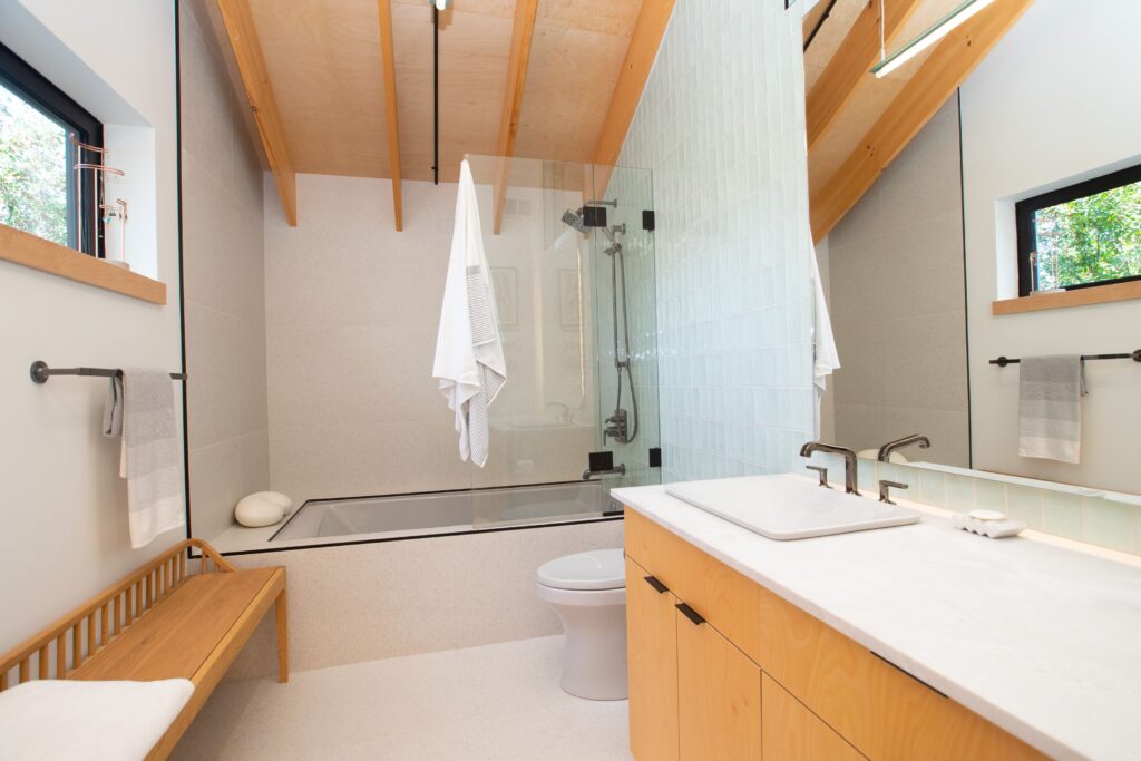 Modern bathroom with wooden accents, a glass-enclosed bathtub and shower, a white sink on a light wood vanity, a bench, white towels, and a slanted wood ceiling with exposed beams.