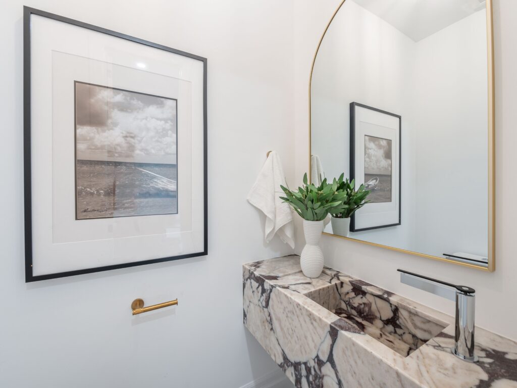 A modern bathroom with a marble sink, a large mirror with a gold frame, a white vase of green leaves, a towel hanging on the wall, and a framed ocean photograph above a gold toilet paper holder.