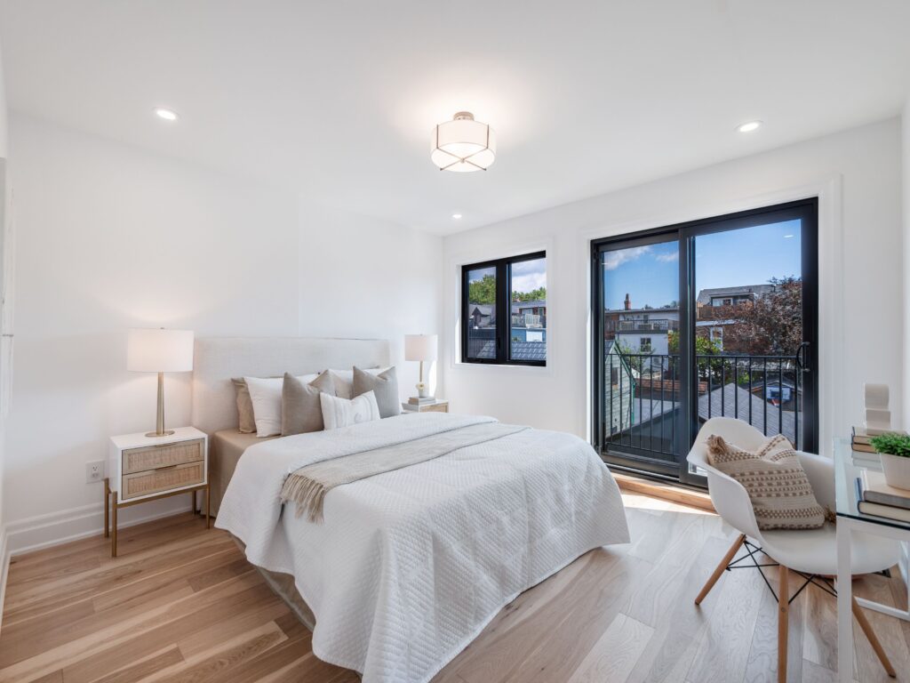 A bright, modern bedroom with a large bed, neutral bedding, two bedside tables with lamps, a small desk and chair, light wood floors, and glass doors leading to a balcony with a city view.