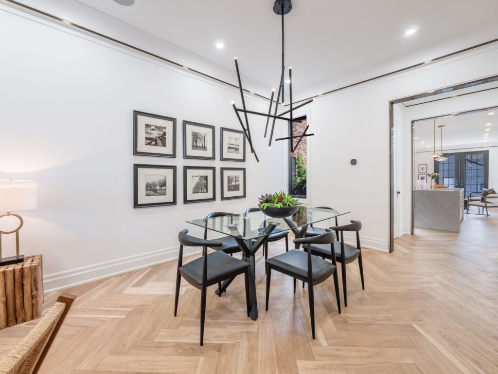 Modern dining area with a glass table, four black chairs, geometric black chandelier, herringbone wood flooring, framed photos on the wall, and an open view to a bright kitchen and living space.