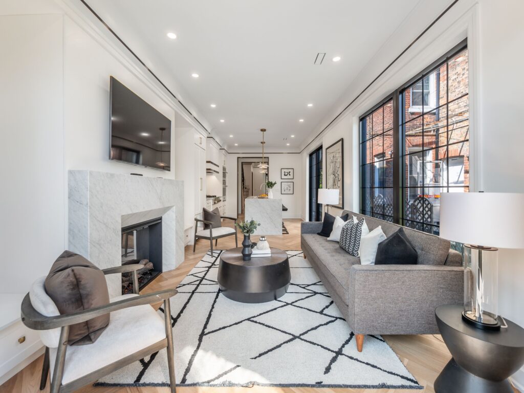 Bright, modern living room with gray sofa, patterned rug, marble fireplace, large wall-mounted TV, armchairs, round coffee table, and large windows letting in natural light. Contemporary decor and neutral tones throughout.