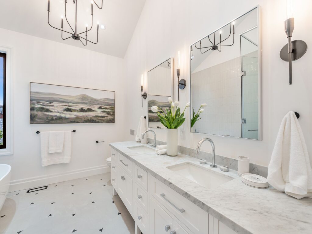 Bright, modern bathroom with double sinks, marble countertop, white cabinets, wall-mounted mirrors, black fixtures, a vase of white tulips, towel holders, and landscape artwork on the wall.