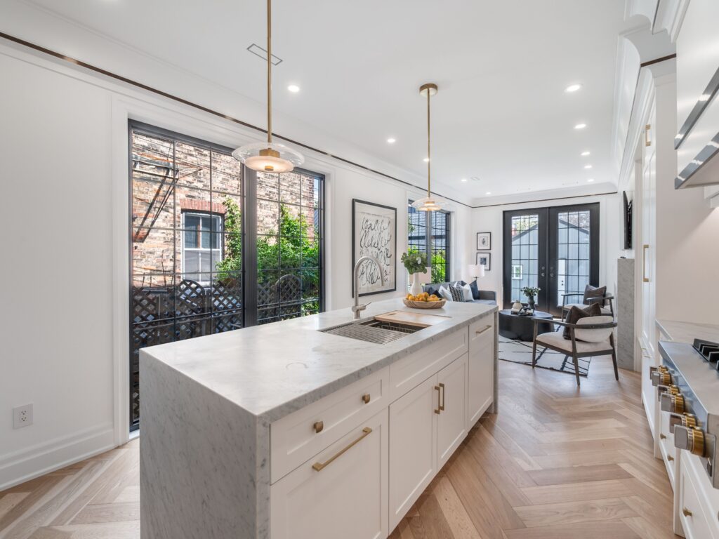 Bright modern kitchen with a marble island, gold accents, and pendant lights, opening to a cozy living area with large windows and French doors, herringbone wood floors, and contemporary decor.