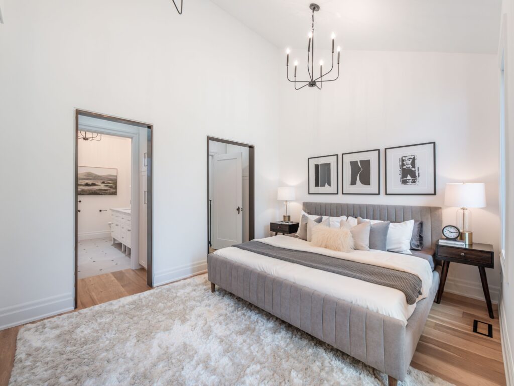 A modern bedroom with a large gray upholstered bed, white bedding, and decorative pillows. The room has hardwood floors, a plush rug, two bedside tables with lamps, abstract wall art, and a view into a bright bathroom.