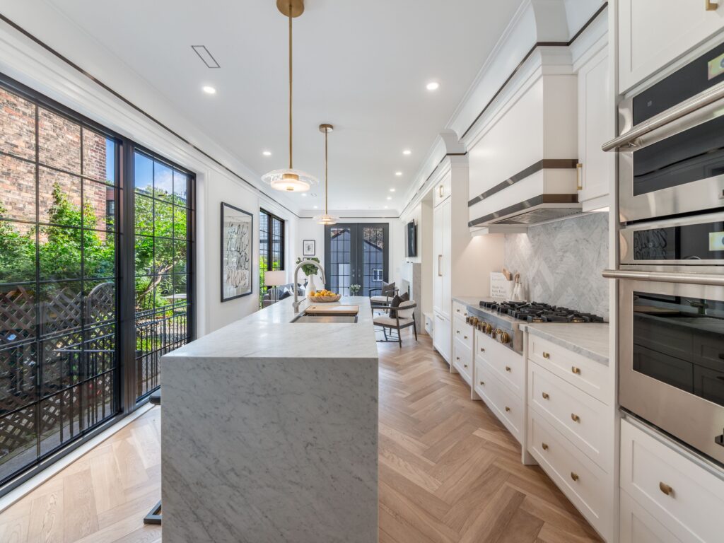 Modern kitchen with white cabinets, marble island, wood herringbone floor, stainless steel appliances, pendant lights, large windows, and a view of a garden patio with greenery outside.