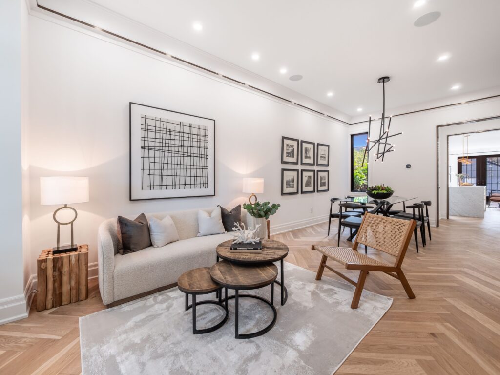 A modern living and dining area featuring a cream sofa, wooden nesting tables, woven chair, abstract wall art, and a large dining table with black chairs. Light wood flooring and neutral tones create an airy, elegant space.