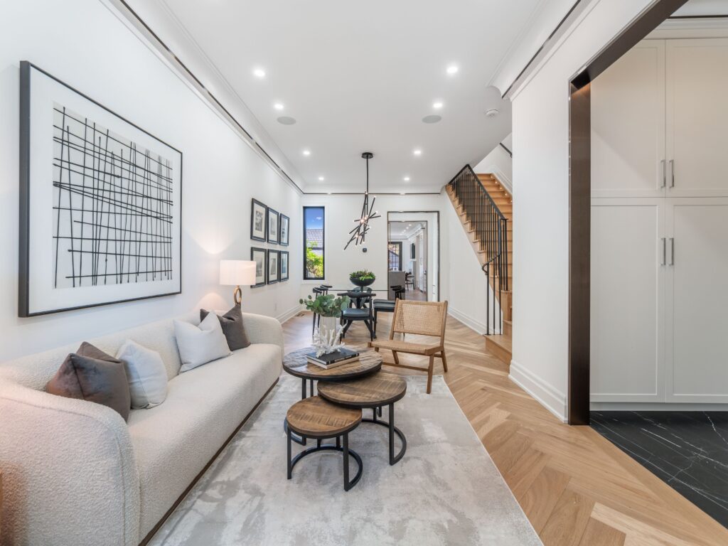 A modern living room with a white sofa, abstract wall art, round coffee tables, and two chairs. The space has wood flooring, a dining area, staircase, and built-in cabinets, with recessed lighting and large windows.