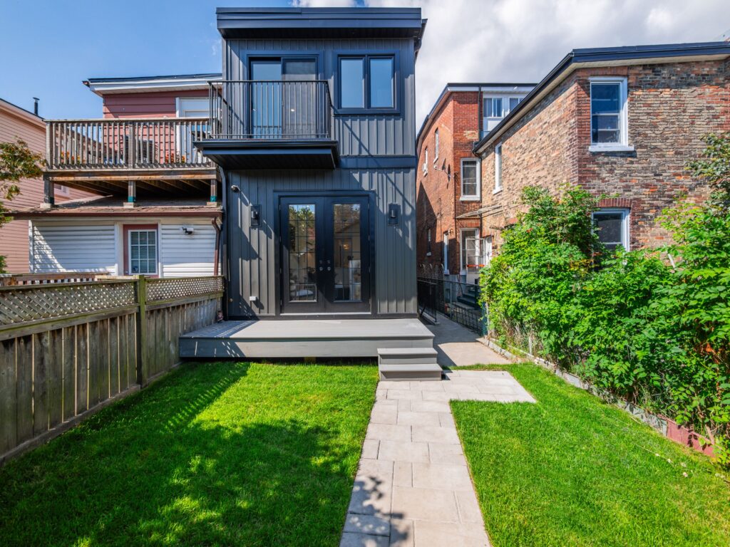 A modern two-story house with dark gray siding and large windows, featuring a small upper balcony and lower patio, a fenced yard, and a stone walkway leading to the back door, surrounded by neighboring brick homes.