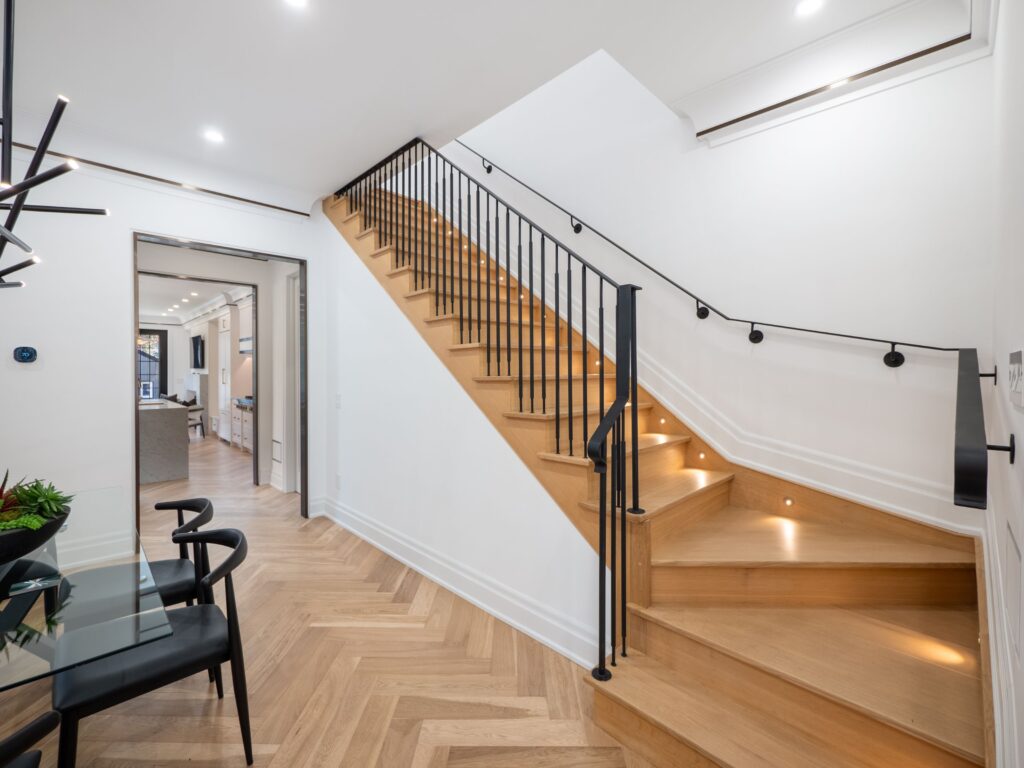 Modern interior with wooden stairs featuring black metal railings, herringbone wood flooring, white walls, and recessed lighting. A dining table and chairs are visible to the left, with a living area in the background.