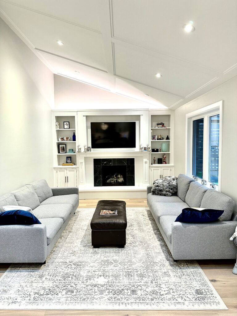 A modern living room with two gray sofas facing each other, a patterned rug, a dark ottoman, and a white fireplace with a large TV above it. Built-in shelves hold decor items, and large windows let in natural light.
