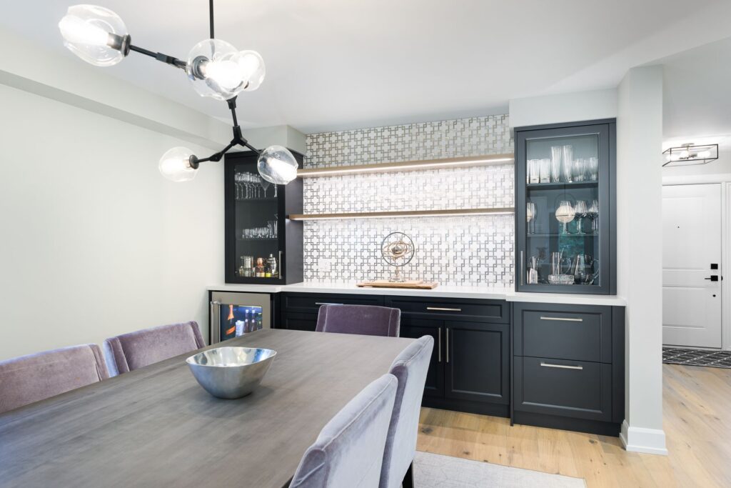Modern dining area with a gray wooden table, velvet chairs, a metallic bowl centerpiece, and a black geometric chandelier. Behind is a built-in bar with glassware, dark cabinets, and patterned tile backsplash.