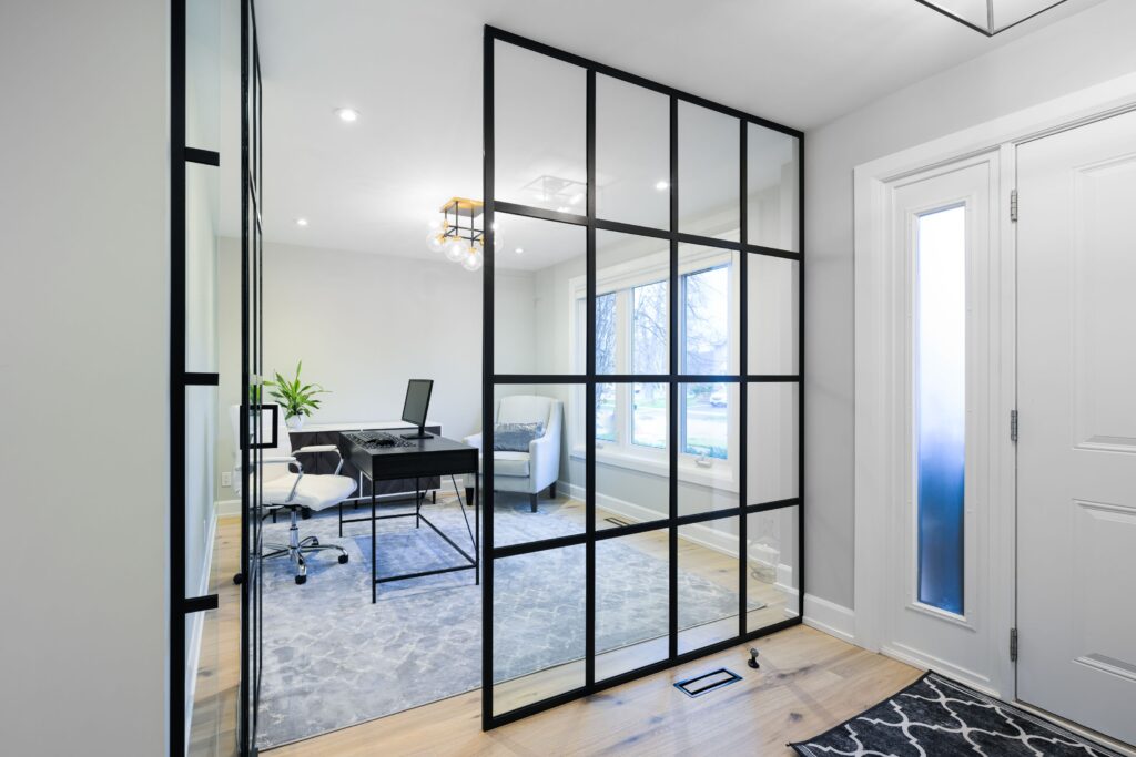 Modern home office with glass partition walls, a black desk with a computer, white chair, an armchair, large window, potted plant, and light wood flooring. The entryway is visible with frosted glass panels on the door.