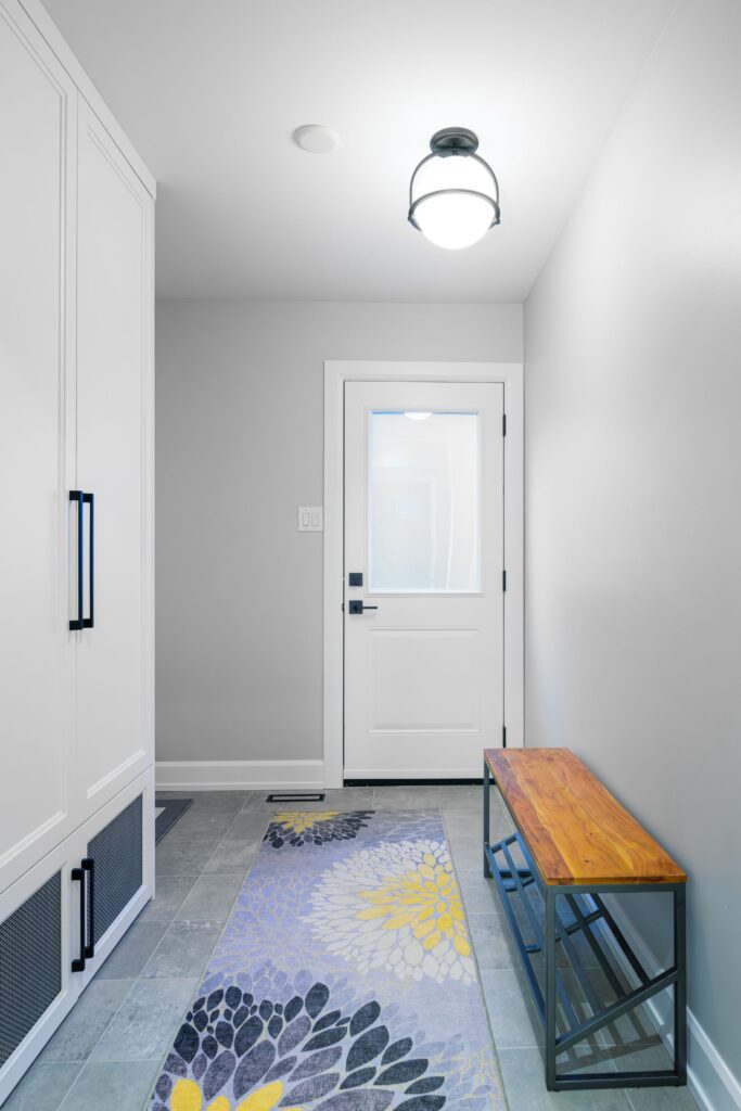 A bright entryway with a white door, gray walls, a patterned rug with yellow and gray floral designs, a wooden bench with metal legs, and white built-in storage cabinets. A ceiling light fixture hangs above.