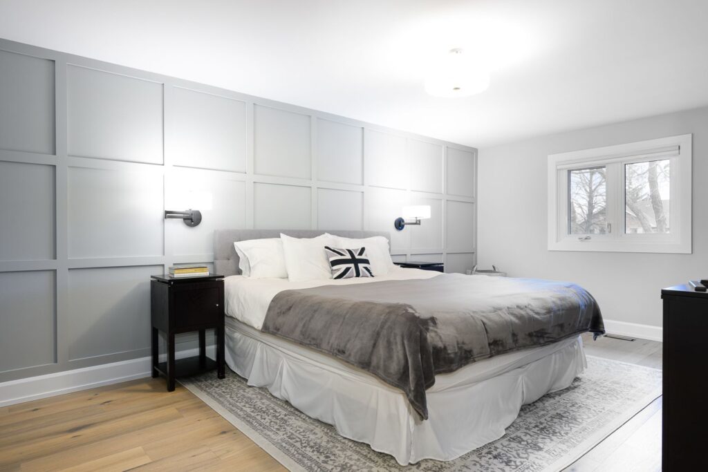 A modern bedroom with light grey paneled walls, a large bed with white and grey bedding, two black nightstands, wall-mounted lamps, a window, and light wooden floors.