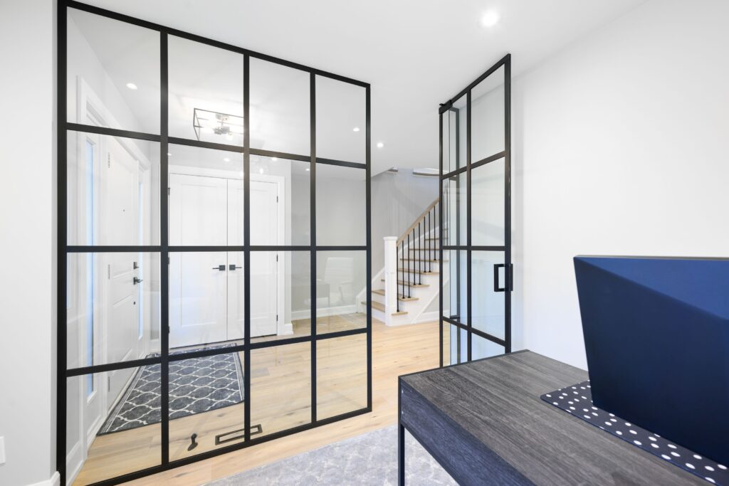 Modern home interior with black-framed glass doors, light wood flooring, white walls, a patterned rug, a staircase, and a dark desk with a blue laptop in the foreground.