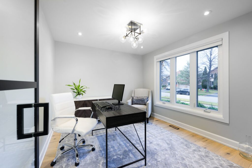 Modern home office with a black desk, white chair, computer monitor, and a potted plant. There is a gray armchair, large window with outside view, light wood floor, and a contemporary ceiling light.