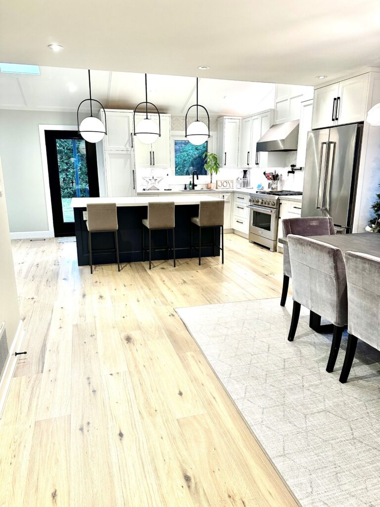 Modern kitchen and dining area with light wood floors, white cabinets, stainless steel appliances, a large island with four beige chairs, pendant lights, and a dining table with gray upholstered chairs on a light rug.