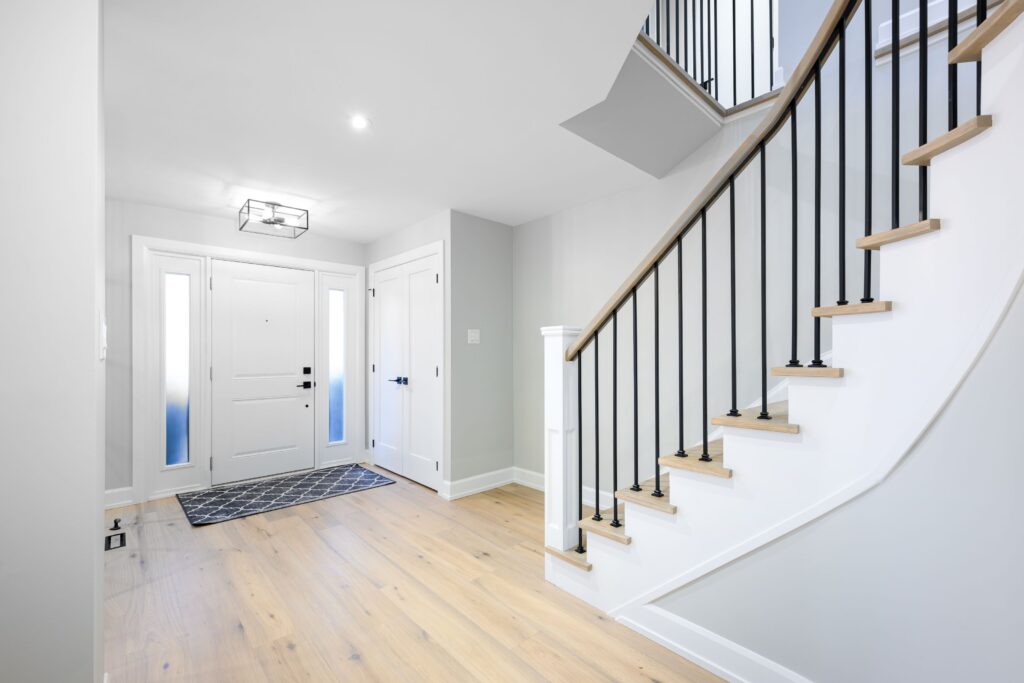 Bright entryway with light wood floors, a white front door with sidelights, a geometric rug, white walls, and a staircase with black metal railings and wooden steps leading to an upper floor.