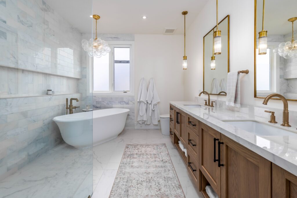 Modern bathroom with marble floors and walls, a freestanding bathtub by a window, a double wooden vanity with two mirrors, gold fixtures, pendant lights, and two white robes hanging on the wall.