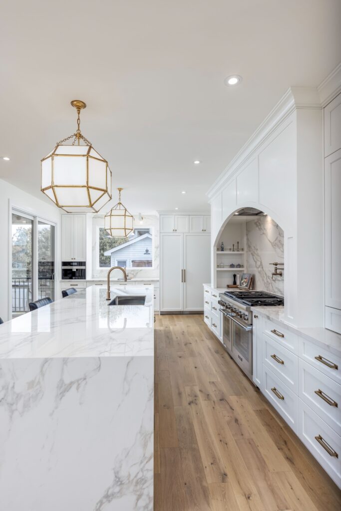 Modern white kitchen with marble countertops, gold pendant lights, white cabinetry with gold handles, stainless steel appliances, and light wood flooring; large windows let in natural light.