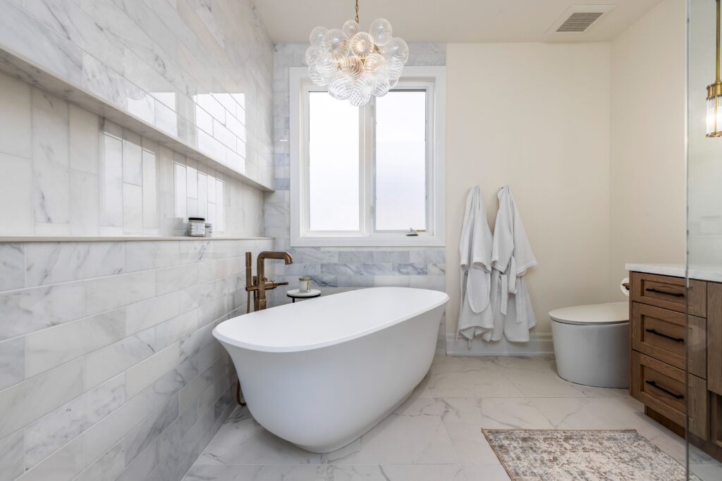 Modern bathroom with a freestanding white tub, marble tile walls and floor, large window, glass bubble chandelier, wooden vanity, two hanging white robes, and a small rug.