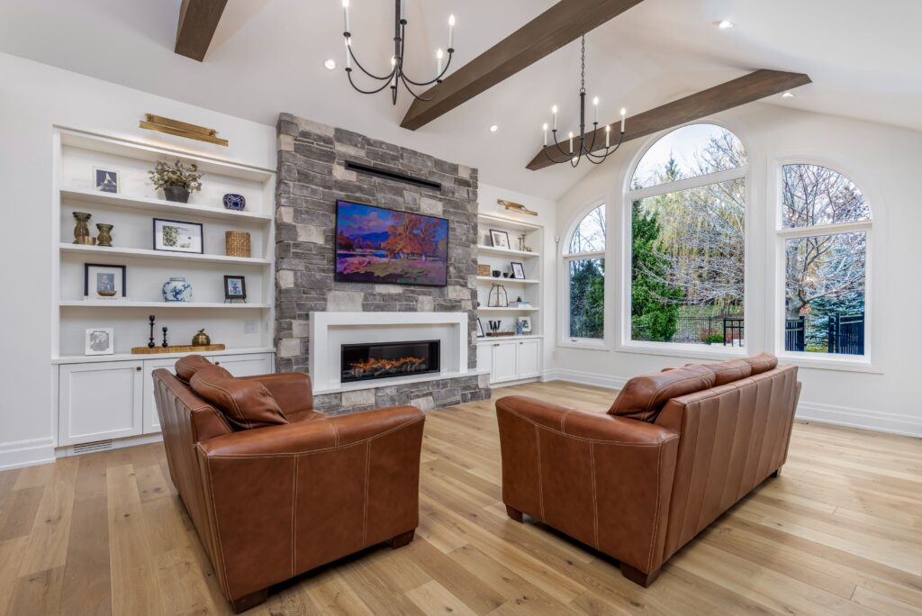 A bright living room with two brown leather sofas facing a stone fireplace and TV, built-in white shelves with decor, wood ceiling beams, and large arched windows showing trees outside.