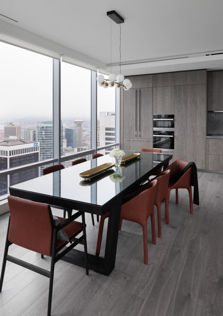 A modern kitchen and dining area with floor-to-ceiling windows, a black dining table, red chairs, light wood cabinets, built-in ovens, and city skyline views in the background.
