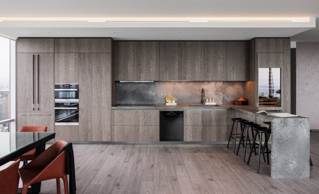 Modern kitchen with gray wood cabinets, integrated appliances, a stone backsplash, a built-in oven, an island with bar stools, and dining chairs with orange cushions. Large windows provide natural light.