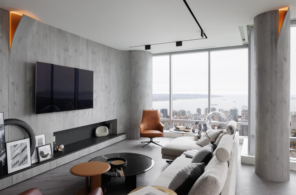 Modern living room with large windows overlooking a city and water view, featuring a wall-mounted TV, gray walls, contemporary furniture including a leather chair and sofa, and minimalist decor.