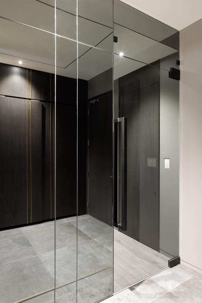 A modern interior with large floor tiles, mirrored glass walls, and dark wood panels. The space is minimalistic, with clean lines and subtle ceiling lights illuminating the sleek, contemporary design.